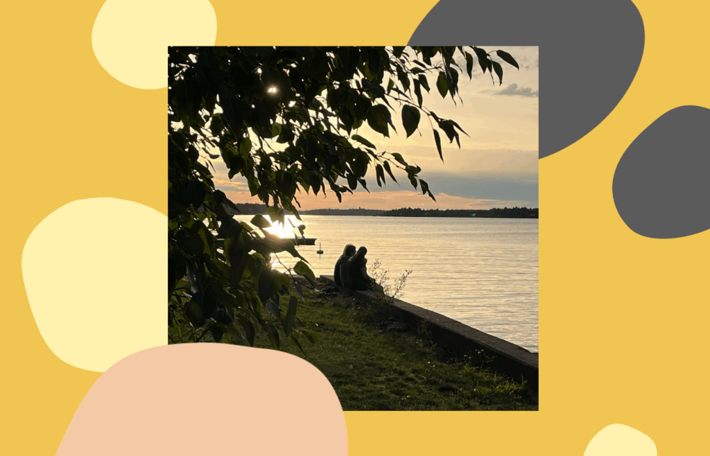 Två ungdomar vid strandkanten tittar ut mot solnedgång över en sjö. I förgrunden löv och i bakgrunden glittrar solen i vattnet.